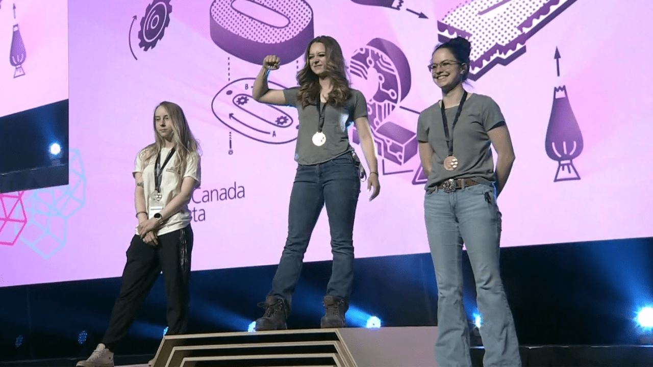 Three women stand on a podium in first, second and third. The first-place winner has her first in the air celebrating. The silver-medal winner stands smiling with her arms crossed behind her; the third-place winner has a slight smile and stands with her right foot forward, leaning back, and her arms are crossed in front of her. 